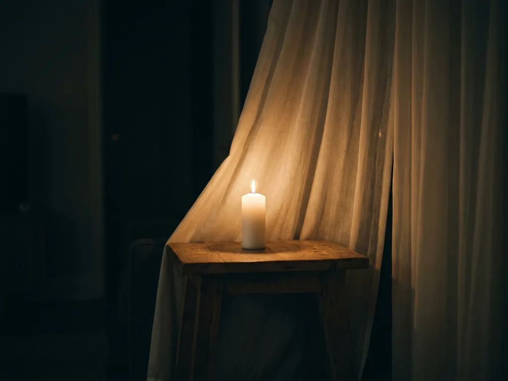 White pillar candle burning on a wooden side table, amber flame dangerously close to sheer curtains in a dim living room.