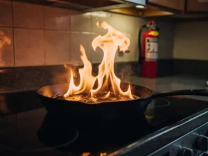 Cast iron pan grease fire with tall orange flames on stovetop, fire extinguisher mounted on kitchen wall in background.