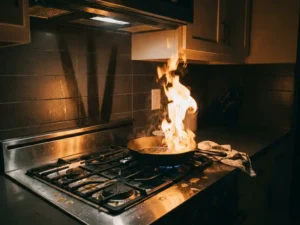 Cast iron pan engulfed in grease fire on gas stove, flames reaching high, kitchen towel dangerously nearby.