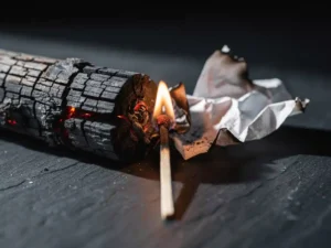 Charred log, crumpled paper, and matchstick forming a triangle on dark slate, single flame burning at their center.