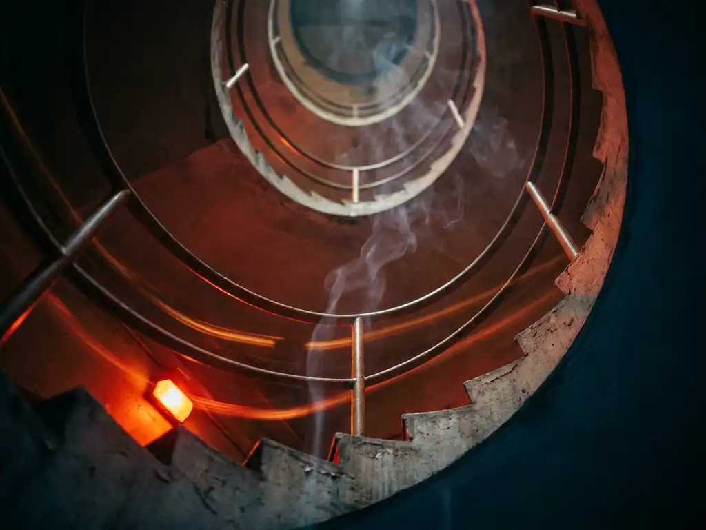 Concrete stairwell viewed from below with emergency red-amber lighting, smoke rising through the shaft, and deep shadows along metal handrails.