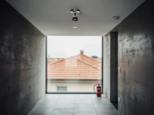 Fire sprinkler head on a concrete apartment corridor ceiling, with a red fire extinguisher on the tiled floor and a suburban house visible through a window.