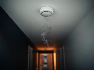 White smoke detector on ceiling with grey smoke rising toward it, warm amber glow from distant doorway suggesting early fire.