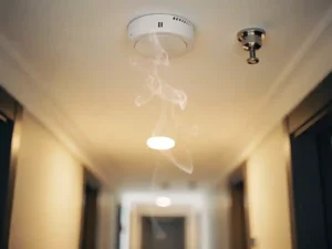 Smoke detector on white ceiling with pale smoke rising toward it, sprinkler head nearby, warm amber interior lighting.